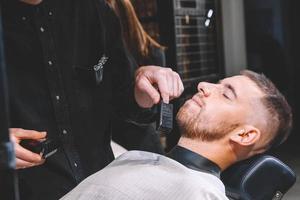 El peluquero masculino afeita la barba del cliente con una recortadora eléctrica. Joven barbudo cortarse el pelo por peluquero en peluquería foto