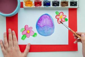 The child's hand draws an Easter egg and flowers in watercolor on a white sheet. photo