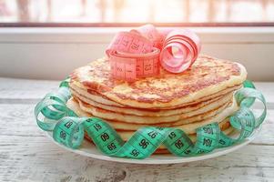 a stack of pancakes on a plate with a centimeter tape on top on a wooden table. the concept of an unhealthy lifestyle. overweight. photo