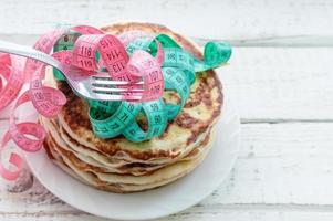 a hand holds a fork with a centimeter-long ribbon over a stack of pancakes on a plate on a wooden table. the concept of an unhealthy lifestyle. overweight. photo