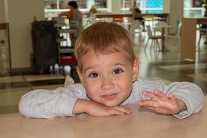 Un niño hermoso y alegre hace muecas en un café en una mesa en el patio de comidas del centro comercial. compras. foto