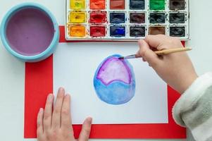 The child's hand draws an Easter egg and flowers in watercolor on a white sheet. photo
