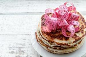 a stack of pancakes on a plate with a centimeter tape on top on a wooden table. the concept of an unhealthy lifestyle. overweight. photo