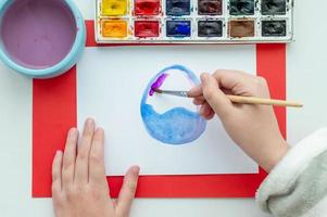 The child's hand draws an Easter egg and flowers in watercolor on a white sheet. photo