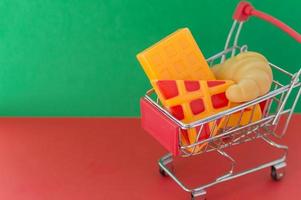 Shopping basket on a red and green background with plastic pizza, croissants and waffles inside. The concept of unnatural products with harmful additives. photo