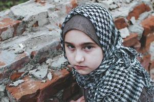 a refugee child in the war, a Muslim girl with a dirty face on the ruins, the concept of peace and war, the child is crying and waiting for help. photo