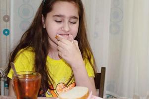 una adolescente está sentada en una mesa en la cocina y toma té con un sándwich. mañana, desayuno, comida chatarra. foto