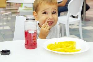 El niño pequeño come papas fritas y bebe jugo, sentado a la mesa en el patio de comidas del centro comercial. comida chatarra. foto