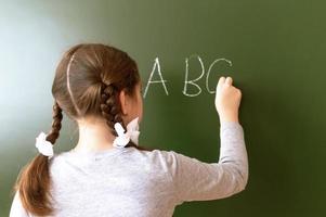 Elementary school girl stands in front of a blackboard and writes with chalk photo