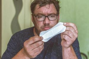 Un hombre con gafas mira una toalla sanitaria femenina con sorpresa. foto