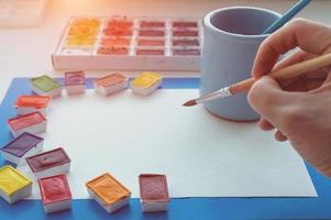 la mano del artista sostiene un pincel sobre una hoja de papel blanca. pinturas de acuarelas, vaso con agua. de cerca. foto