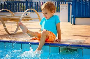 A small smiling boy sits on the edge of the pool, splashing with his feet in the water. Tourism, travel. photo