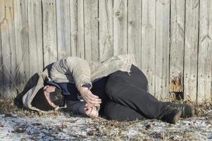 Homeless. At the wooden fence lies the body of a man in dirty and torn clothes with a medical mask on his face. photo
