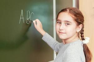 Niña de la escuela primaria se para frente a una pizarra y escribe con tiza foto