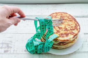 a hand holds a fork with a centimeter-long ribbon over a stack of pancakes on a plate on a wooden table. the concept of an unhealthy lifestyle. overweight. photo
