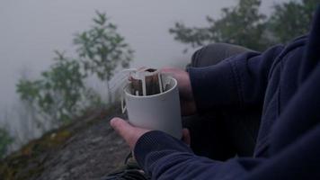 Primer plano de una taza con café o té caliente en la mano del viajero en el pico de la montaña con una espesa niebla en las primeras horas de la mañana.