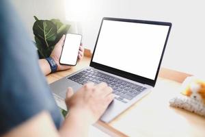 Mockup image of man's hands holding white mobile phone and laptop with blank screen on thigh and coffee cup in cafe photo