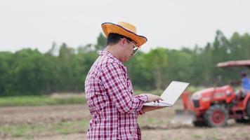 Aziatische boer staat en werkt op laptop op boerderij. rondkijken en een plantenprobleem oplossen door op de laptop te typen en de tractor op de achtergrond te scheren. landbouw werken. agrarisch concept