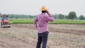 granjero asiático con smartphone, hablando por teléfono celular en un gran campo mientras tractor desgarradoras en segundo plano. trabajo agrícola. concepto agrícola