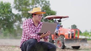 granjero asiático sentado y trabajando en una computadora portátil en la granja, tomando una nota de problema de plantas en la computadora portátil y dejando que el tractor se ponga en marcha primero. trabajo agrícola. concepto agrícola video