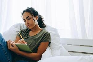 Mujer africana meditativa escribiendo algo en el bloc de notas en la cama foto