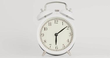 despertador branco com longos ponteiros negros indicando o tempo 6 horas e 10 minutos em um fundo branco.