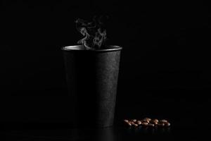 un vaso negro de café caliente con granos de café esparcidos sobre un fondo negro. de cerca foto