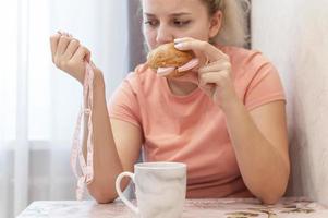 una niña con sobrepeso está comiendo un sándwich y sosteniendo una cinta de un centímetro de largo. comida chatarra. enfoque selectivo, grano de película.