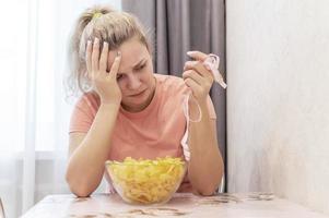 An overweight girl is crying, eating potato chips and holding a centimeter-long ribbon. Junk food. Selective focus, film grain. photo