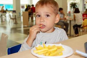 little child eating french fries sitting at a table on the food court of the mall. Unhealthy food. photo