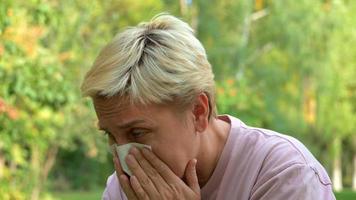 una niña con cabello rubio y un corte de pelo corto estornuda por alergias y se limpia la nariz con una servilleta en el contexto de la naturaleza verde