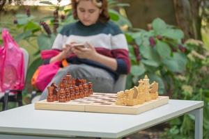 una adolescente con un corte de pelo corto está sentada frente al ajedrez y jugando al teléfono en la naturaleza