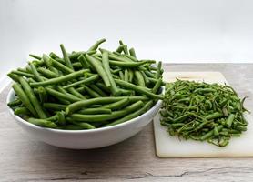 Raw green beans in a white bowl on wooden table with scraps on the side photo
