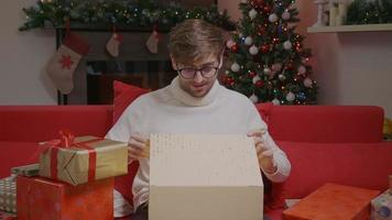 Young man is opening a gift box, light shines on face, Christmas lights on the background. video