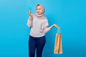 Portrait of attractive asian woman holding shopping bag and showing credit card over blue background photo