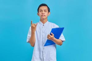 Portrait of smiling male nurse holding clipboard and showing rock gestureon blue background photo