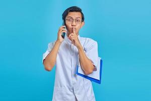 Male nurse talking on mobile holding clipboard and making silent gesture on yellow background photo