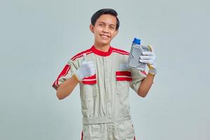 Retrato de alegre apuesto hombre vestido con uniforme de mecánico sosteniendo una botella de plástico de aceite de motor y mostrando aprobación con el pulgar hacia arriba foto