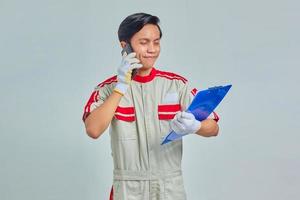 Portrait of smiling young Asian mechanic talking on phone and holding clipboard isolated over gray background photo