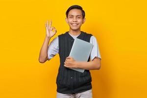 Handsome Asian man holding laptop and showing gesturing okay sign over  yellow background photo