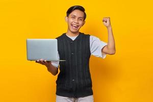 Excited Asian man holding laptop and celebrating victory smiling over yellow background photo