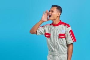 Retrato de sonriente joven mecánico asiático gritando y gritando fuerte hacia los lados con la mano en la boca sobre fondo azul. foto