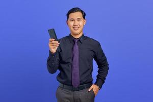 Cheerful young handsome businessman holding smartphone and looking at camera on purple background photo