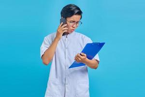 Retrato de enfermero sonriendo y hablando por teléfono inteligente mientras sostiene el portapapeles aislado sobre fondo azul. foto