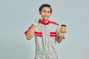 Feliz joven mecánico hablando por teléfono inteligente y sosteniendo café sobre fondo gris foto
