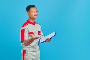 Portrait of a cheerful young mechanic holding clipboard and pointing with palm at camera isolated on blue background