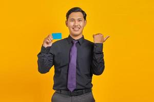 Sonriente empresario asiático sosteniendo una tarjeta de crédito mientras señala con el dedo en el espacio de la copia en amarillo foto