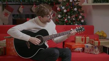 Man playing guitar singing Christmas songs sitting near a decorated home.