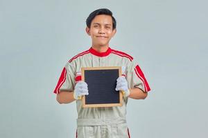 Retrato de sonriente joven mecánico vistiendo uniforme mostrando placa en blanco sobre fondo gris foto