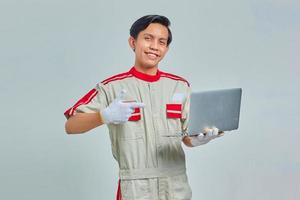 Smiling handsome young mechanic man wearing uniform pointing to laptop on gray background photo